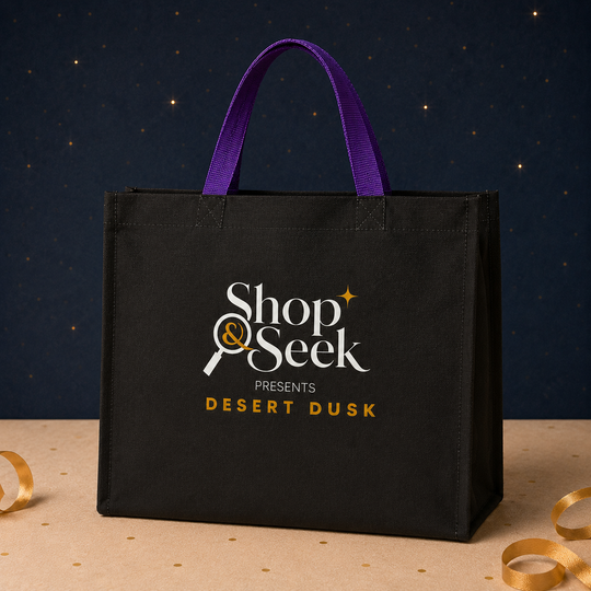 A black fabric tote bag with purple handles stands on a beige surface against a dark navy starry backdrop. The bag has a printed logo reading &